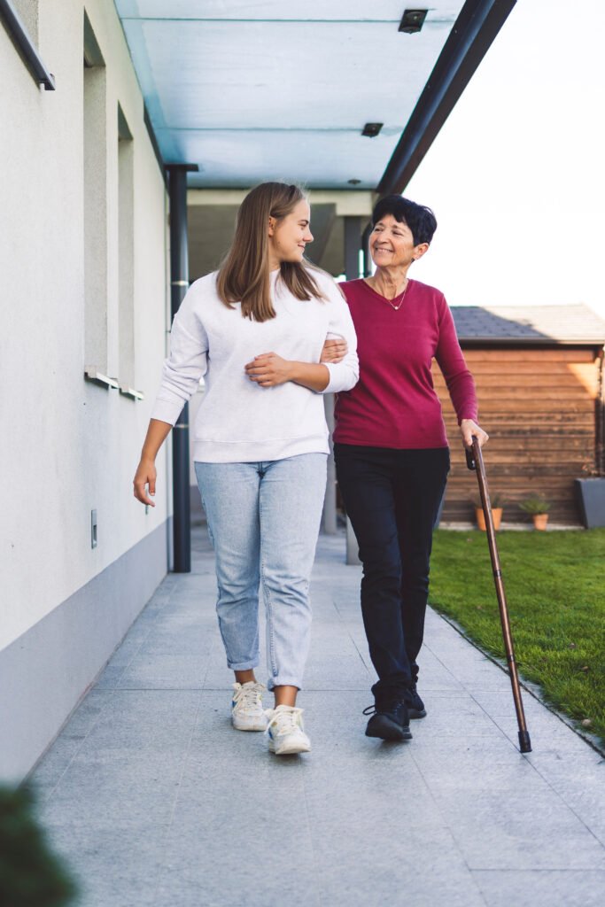 Caregiver walking with elderly woman using cane showing emotional support at home