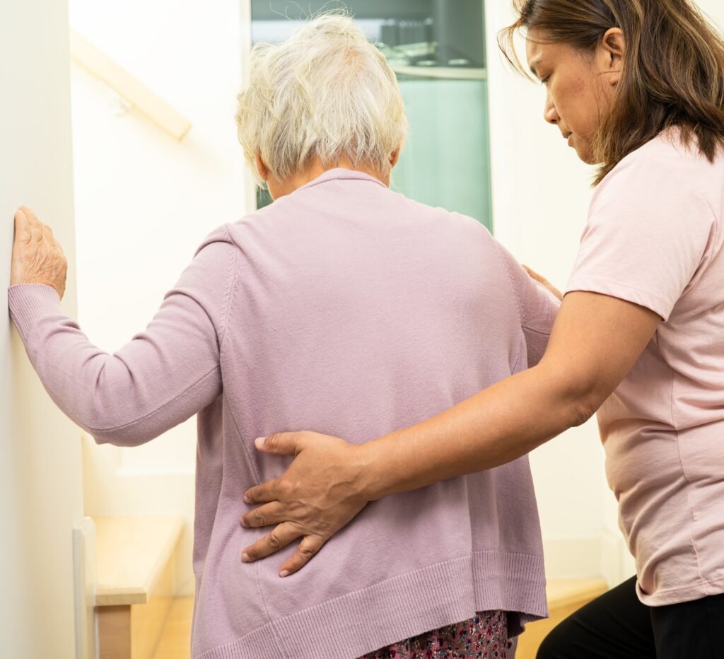 Older woman with dementia using a wall for support while walking as a caregiver stands nearby to assist with early mobility changes.